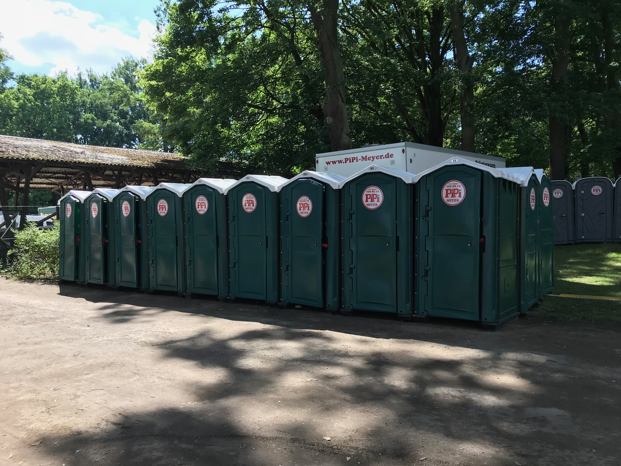 Eine Reihe grüner mobiler Toilettenkabinen von Pipi Meyer, aufgestellt im Freien unter Bäumen, mit einem mobilen Toilettenwagen im Hintergrund.