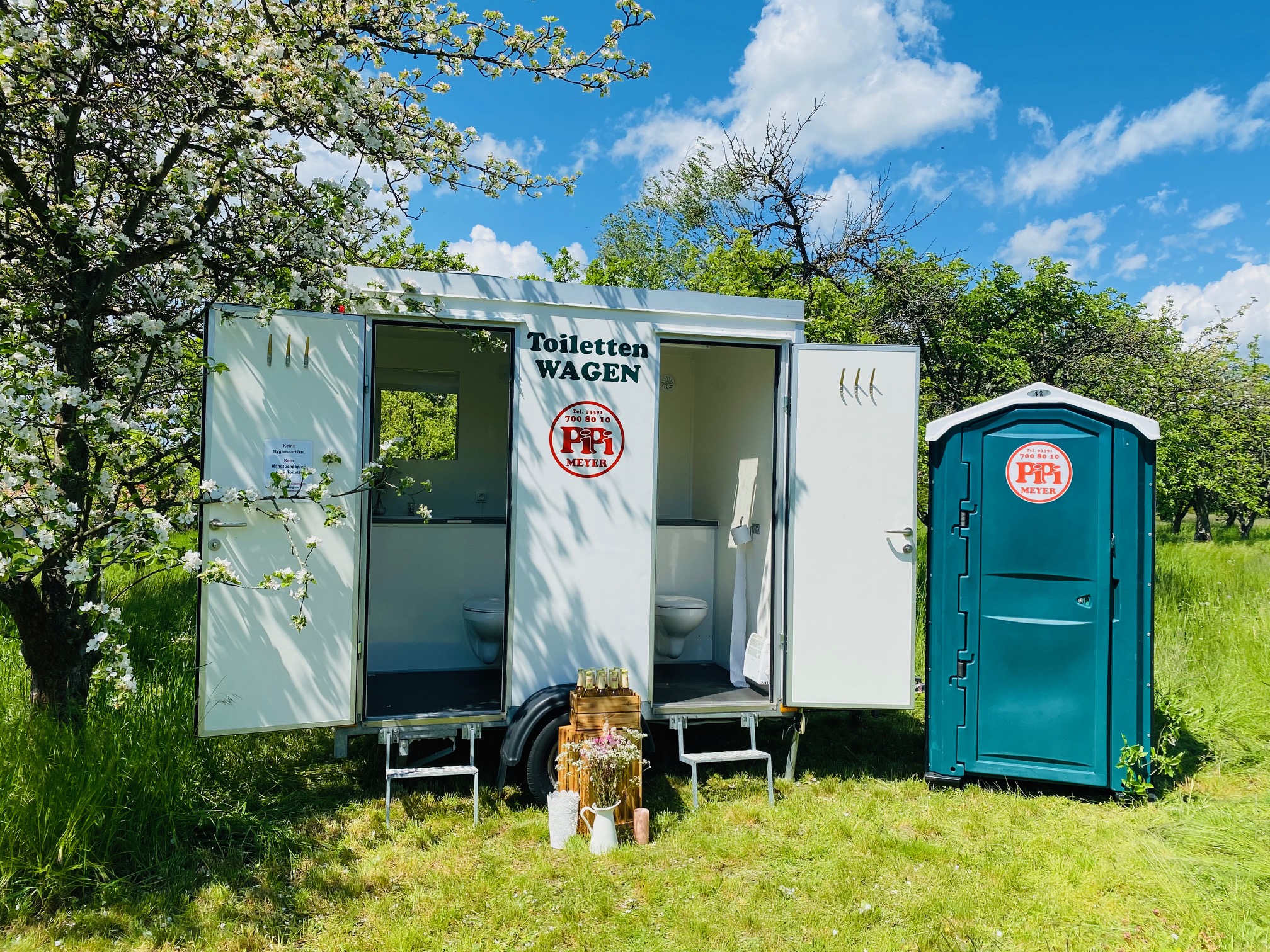 Ein offener mobiler Toilettenwagen und eine grüne mobile Toilettenkabine von Pipi Meyer stehen in einer blühenden Wiese unter Bäumen, mit blauen Himmel und weißen Wolken im Hintergrund.