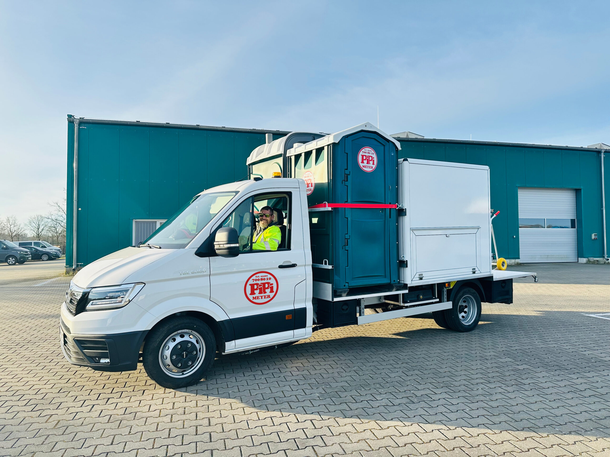 Servicemitarbeiter für Baustellentoiletten sitz im Servicewagen und schaut aus dem Fenster. Auf der Pritsche stehen zwei Baustellentoiletten.